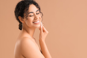 Young African-American woman removing makeup with cotton pad on beige background, closeup