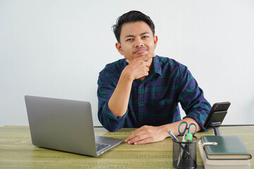Confused young asian man in blue shirt sit work at wooden desk with pc laptop isolated on white background. Achievement business career lifestyle concept. Put hand prop up on chin © DMH