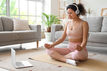 Sporty African-American woman with laptop and headphones meditating on yoga mat at home