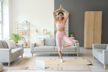 Sporty African-American woman with laptop and headphones practicing yoga at home