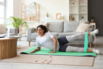 Sporty African-American woman with laptop and resistance band exercising on yoga mat at home