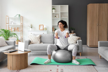 Sporty African-American woman with laptop, dumbbells and fitball exercising on yoga mat at home