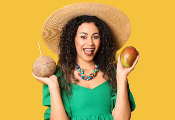 Beautiful African-American woman with fresh mango and coconut on yellow background