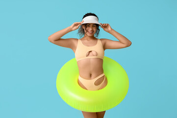 Beautiful young African-American woman in stylish beige swimsuit with inflatable ring on blue...