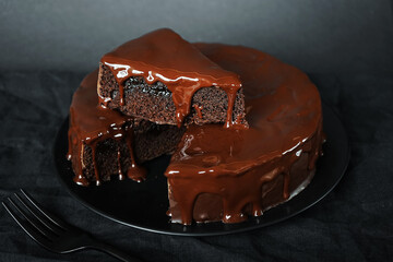 Chocolate cake with chocolate icing on a black plate next to a fork on a dark background