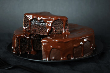 Chocolate cake with chocolate icing on a black plate next to a fork on a dark background