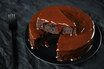 Chocolate cake with chocolate icing on a black plate next to a fork on a dark background