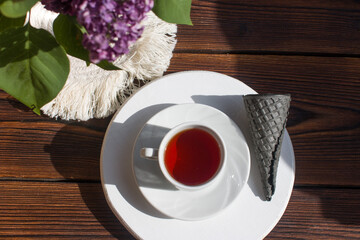 red tea in a mug on a saucer with a waffle black horn and a bouquet of lilacs. Summer hot drink