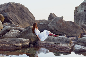 Serene woman in white dress sitting on rocks by water, dipping her feet in the cool water