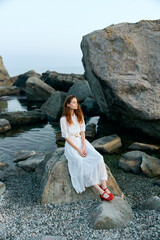 Woman in white dress with red shoes sitting on rock by ocean, contemplating the vast horizon