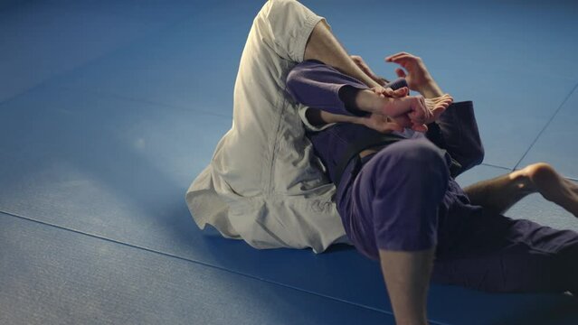 Martial arts athlete in white uniform pulling and throwing his opponent down on mat and doing chokehold during jujutsu practice