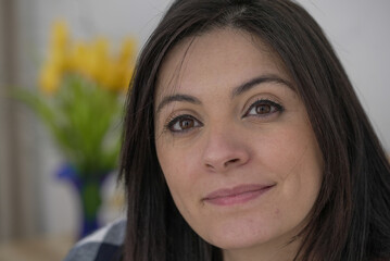 Obraz premium Woman with a thoughtful expression, close-up of face, subtle smile, dark hair, natural beauty, wearing a checkered shirt, indoor setting, blurred yellow flowers in background