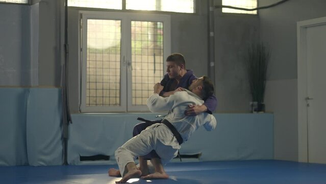 Tilt down shot of young barefoot male fighter in white uniform practicing jujitsu takedown and lockdown with partner in gym