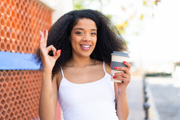 Young African American woman holding a take away coffee at outdoors showing ok sign with fingers