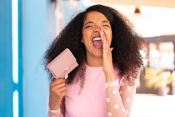 Young African American woman holding a wallet at outdoors shouting with mouth wide open
