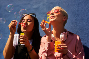 Women, blowing bubbles and fun at wall with urban fashion, streetwear and nostalgia in city. Girl...