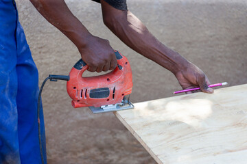 african man using a jigsaw to cut a plank of wood, holding a pencil to mark