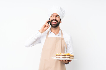 Young man holding muffin cake over isolated white background keeping a conversation with the mobile...