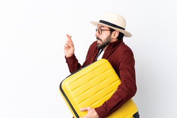 Traveler man man with beard holding a suitcase over isolated white background with fingers crossing...