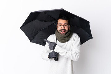 Caucasian handsome man with beard holding an umbrella over isolated white wall surprised and...
