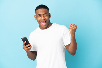Young latin man isolated on blue background using mobile phone and doing victory gesture