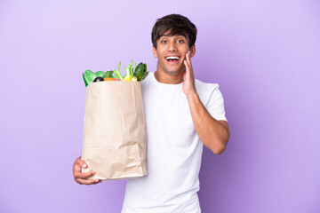 Young man holding a grocery shopping bag isolated on purple background with surprise and shocked facial expression