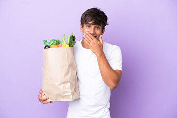 Young man holding a grocery shopping bag isolated on purple background happy and smiling covering...