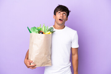 Young man holding a grocery shopping bag isolated on purple background looking up and with...