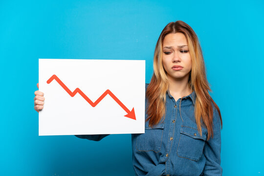 Teenager girl over isolated blue background holding a sign with a decreasing statistics arrow symbol with sad expression