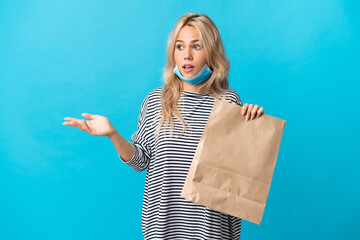 Young Russian woman holding a grocery shopping bag isolated on blue background with surprise...