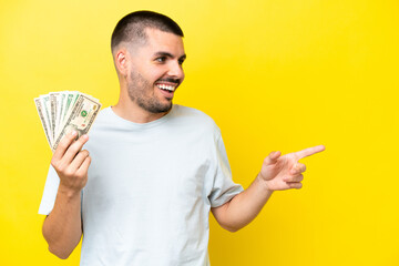 Young caucasian man taking a lot of money isolated on yellow background pointing finger to the side...