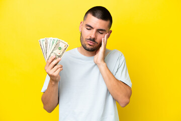 Young caucasian man taking a lot of money isolated on yellow background with headache