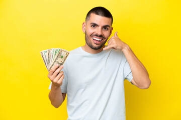 Young caucasian man taking a lot of money isolated on yellow background making phone gesture. Call...