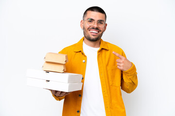 Young caucasian man holding pizzas and burgers isolated on background giving a thumbs up gesture