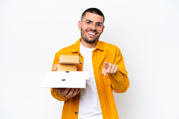 Young caucasian man holding pizzas and burgers isolated on background surprised and pointing front
