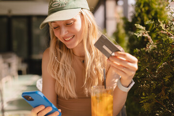 Happy young blonde woman with cap customer shopper holding credit card using cell phone mobile app buying fashion clothes paying online make purchase in ecommerce digital store on smartphone in cafe.