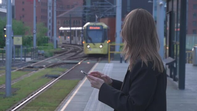 Woman in business casual clothes is standing at a public transport stop looking at her phone and a tram arriving at the stop