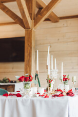 The table for a romantic dinner is decorated with red roses in vases and candles, as well as petals.