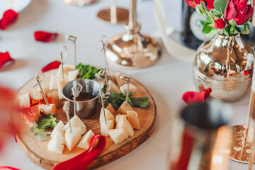 The table for a romantic dinner with cheese plate is decorated with red roses in vases and candles, as well as petals.