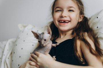 a smiling child without 1 baby tooth sitting on the bed at home, hugging and stroking his kitten