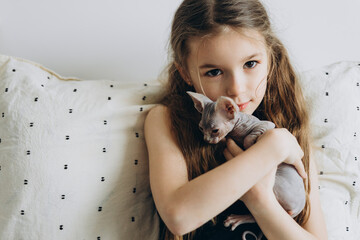 a smiling child without 1 baby tooth sitting on the bed at home, hugging and stroking his kitten