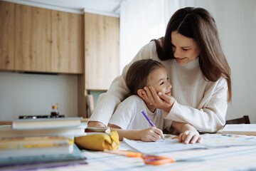 A mother hugs and kisses her daughter while she does her school homework at home at the kitchen table