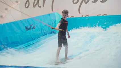 Water sport activity, balance concept. Little athlete enjoying indoor surfing. Teenager rides board on waves on simulator in sports complex. Surfing coach and student in session on wave simulator.