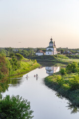 Summer landscape in Suzdal. Church of Elijah the Prophet on the bank of the river Kamenka. Tourists on SUP boards float on the river. Russia, Vladimir region, Golden Ring of Russia