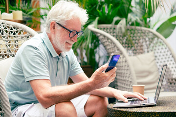 Portrait of smiling relaxed senior man using mobile phone and laptop sitting on armchair. Old generation people and new technologies