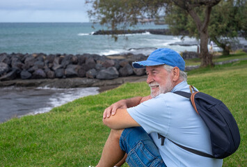 Smiling senior man sitting on the grass at the beach in a cloudy day enjoying free time vacation or...