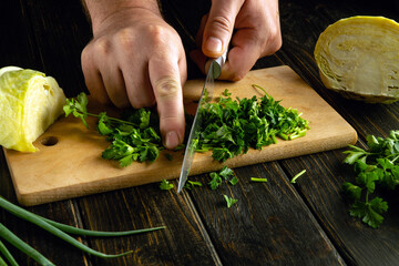 A man hands use a knife to chop fresh green parsley on a cutting board to prepare a vegetable salad...
