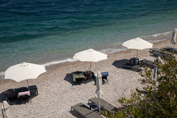 White umbrellas, sunbeds on sundy pebble beach with turquoise crystal clear water in summer morning.
