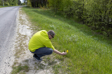 A man in a green jacket kneels on the side of a country road, looking at a patch of wildflowers...