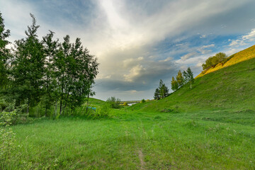 Obraz premium A dirt path winds through lush, green hills, leading toward a distant, shimmering body of water under a cloudy spring sky.